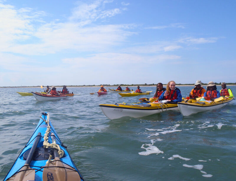 A group of people are kayaking on a sunny day. They are wearing life jackets and paddling in the water. The sky is blue with some clouds. The water is calm and there are some small waves. There are nine kayaks visible in the image. The kayaks are of different colors, including blue, yellow, and white.